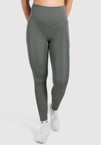 Legginsy w kolorze oliwkowym z wysokim stanem, wykonane z gładkiego, elastycznego materiału. Posiadają płaskie szwy i boczne kieszenie. Noszone z białymi butami sportowymi.