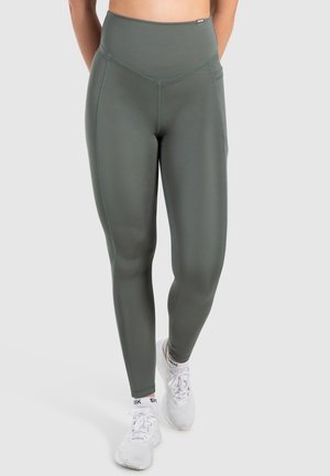 Leggings de tiro alto en color verde oliva, fabricadas con un material suave y elástico. Presenta costuras planas y bolsillos laterales. Combinados con zapatillas deportivas blancas.