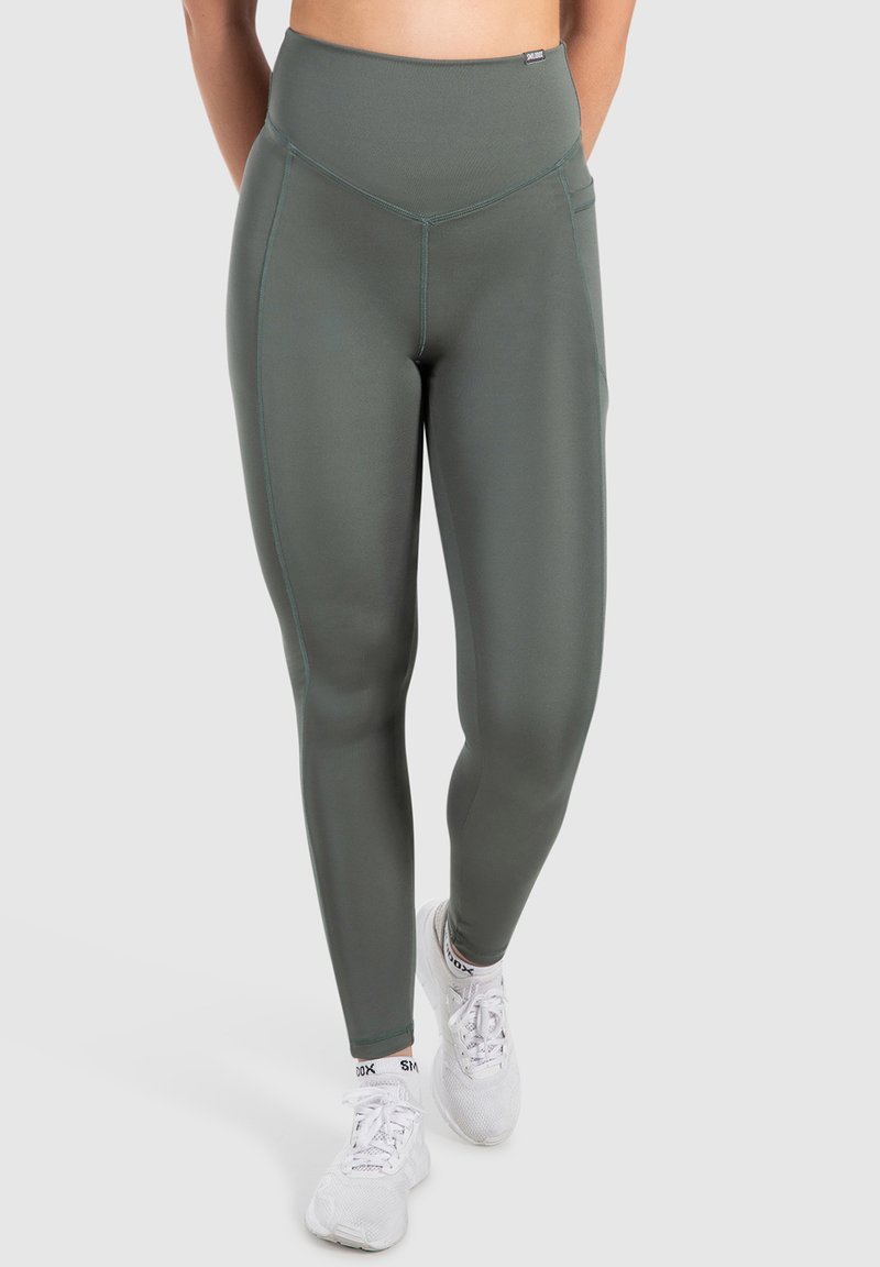 Höga, olivgröna leggings tillverkade av mjukt, stretchigt material. Har platta sömmar och sidofickor. Bärs med vita sneakers.