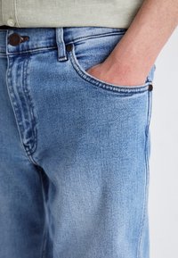 Jean en denim bleu clair avec une coupe décontractée, coutures visibles et une poche sur le côté droit. Le matériau présente une texture lisse.