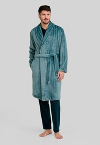 Albornoz de terciopelo turquesa con rayas verticales, cuello chal, cinturilla con hebilla y bolsillos laterales, usado sobre pantalones verde oscuro; textura suave.