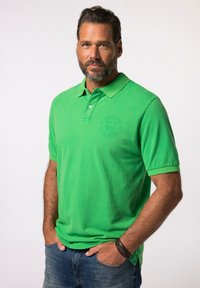 Camisa polo de algodón verde con collar doblado, tapeta de dos botones y logo bordado en el lado izquierdo del pecho, combinada con jeans azules.