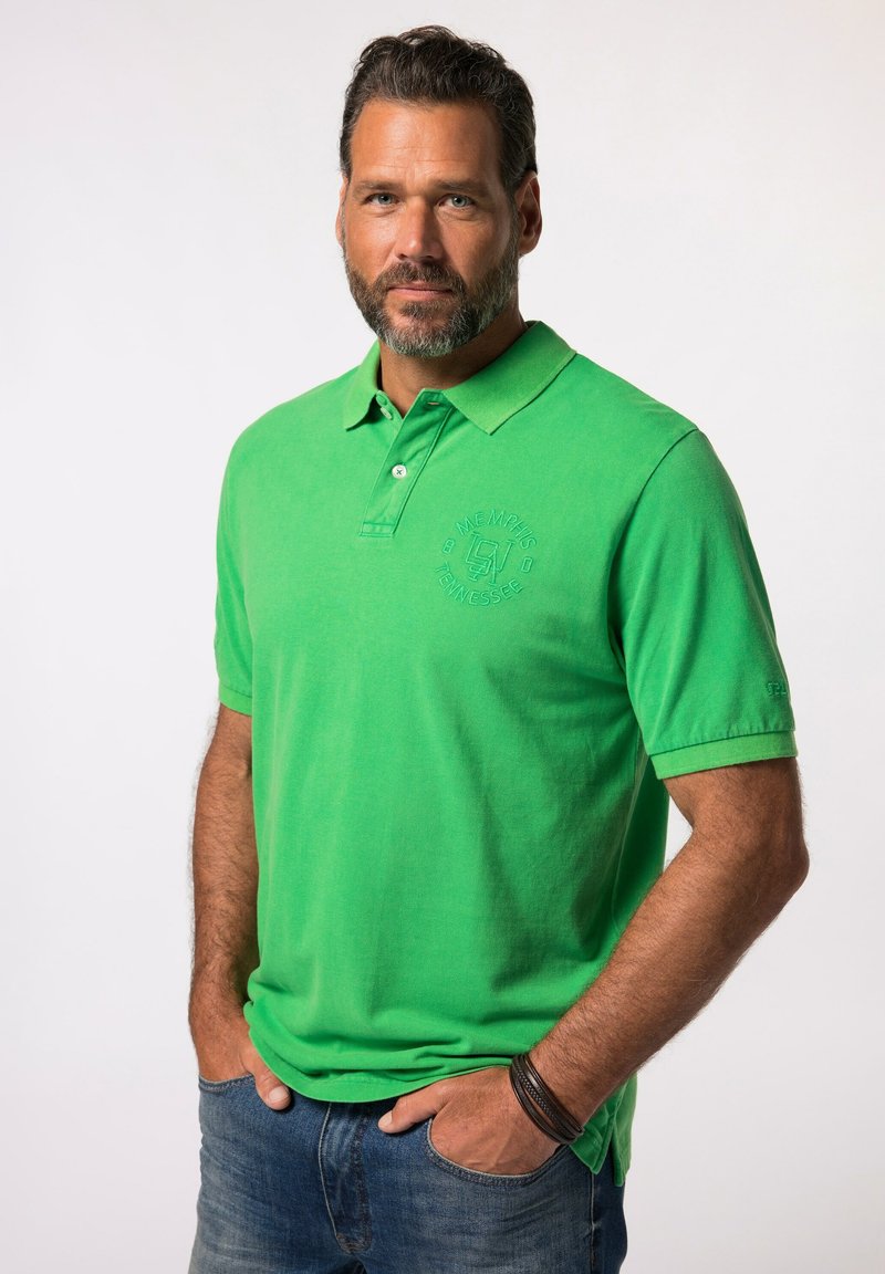 Camisa polo de algodón verde con collar doblado, tapeta de dos botones y logo bordado en el lado izquierdo del pecho, combinada con jeans azules.