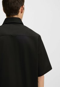Camicia nera a maniche corte con colletto, caratterizzata da una texture di tessuto liscia e un distintivo dettaglio yoke sul retro.