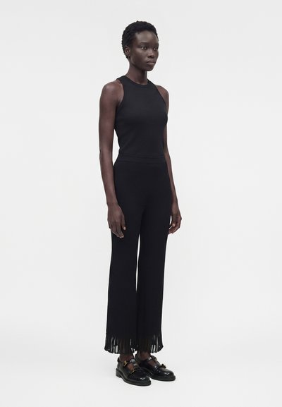 Γυναίκα όρθια, φοράει μαύρο αμάνικο τοπ, μαύρο φαρδύ παντελόνι με φουντίτσες στο τελείωμα και μαύρα γυαλιστερά μοκασίνια.