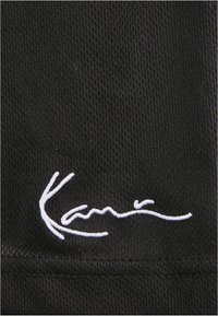Tissu noir à texture côtelée avec le texte brodé en blanc "Kari" dans un style cursif. Aucun motif ou élément matériel notable présent.