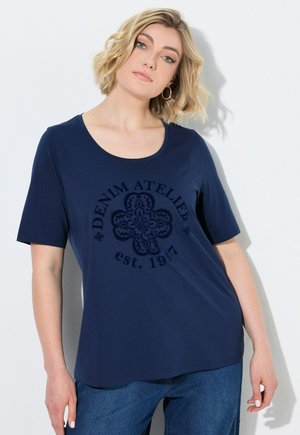 Vrouw met kort blond haar die een donkerblauw T-shirt met de tekst "Denim Atelier est. 1977" en een blauwe spijkerbroek draagt, staand tegen een witte achtergrond.