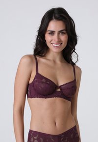 Reggiseno in pizzo bordeaux con stecche, motivo floreale, bordi ondulati e spalline regolabili. Presenta un piccolo dettaglio a fiocco centrale.