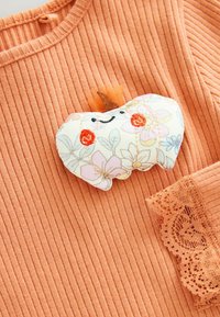 Weiche Stoff-Pumpkin-Brosche mit floralem Muster, orangefarbene Wangen und lächelndes Gesicht auf einem gerippten, orangefarbenen Pullover mit Spitzenbesatz.