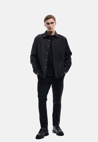 Dunkelgraue Wollmischung Jacke mit Kragen, Knopfverschluss und zwei seitlichen Taschen, kombiniert mit einem schwarzen Hemd und schwarzen Hosen. Schwarze Schuhe runden den Look ab.