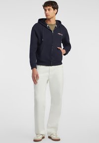 Dunkelblaue Zip-Hoodie mit einer Vorderentasche, Kordelzügen und Logodetail auf der Brust, kombiniert mit weißen Hosen und lässigen Sneakers.