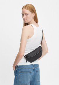 Czarna skórzana nerka o gładkiej fakturze i zaokrąglonym kształcie, z zamkiem błyskawicznym na górze i regulowanym paskiem, noszona na ciele.