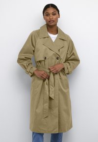 Khakifarbener Trenchcoat mit einem doppelt knöpfbaren Design, großem Kragen, vorderen Taschen und einem gebundenen Gürtel, kombiniert mit einem weißen Hemd und blauen Jeans.