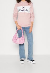 Žena v růžové mikině Hollister, modrých džínách a černobílých teniskách, držící malou růžovou tašku s řasením za její ucho.