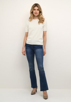 Hvid kortærmet sweater med teksturerede bobler, parret med mørkeblå jeans med en flagrende design og beige højhælede sko.