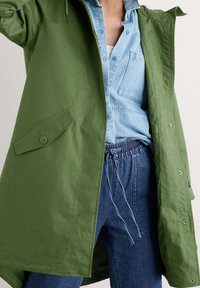 Grön parkas med huva och knappstängning framtill, med sidofickor, över en ljusblå denimskjorta och blå byxor med elastiskt midjeband.