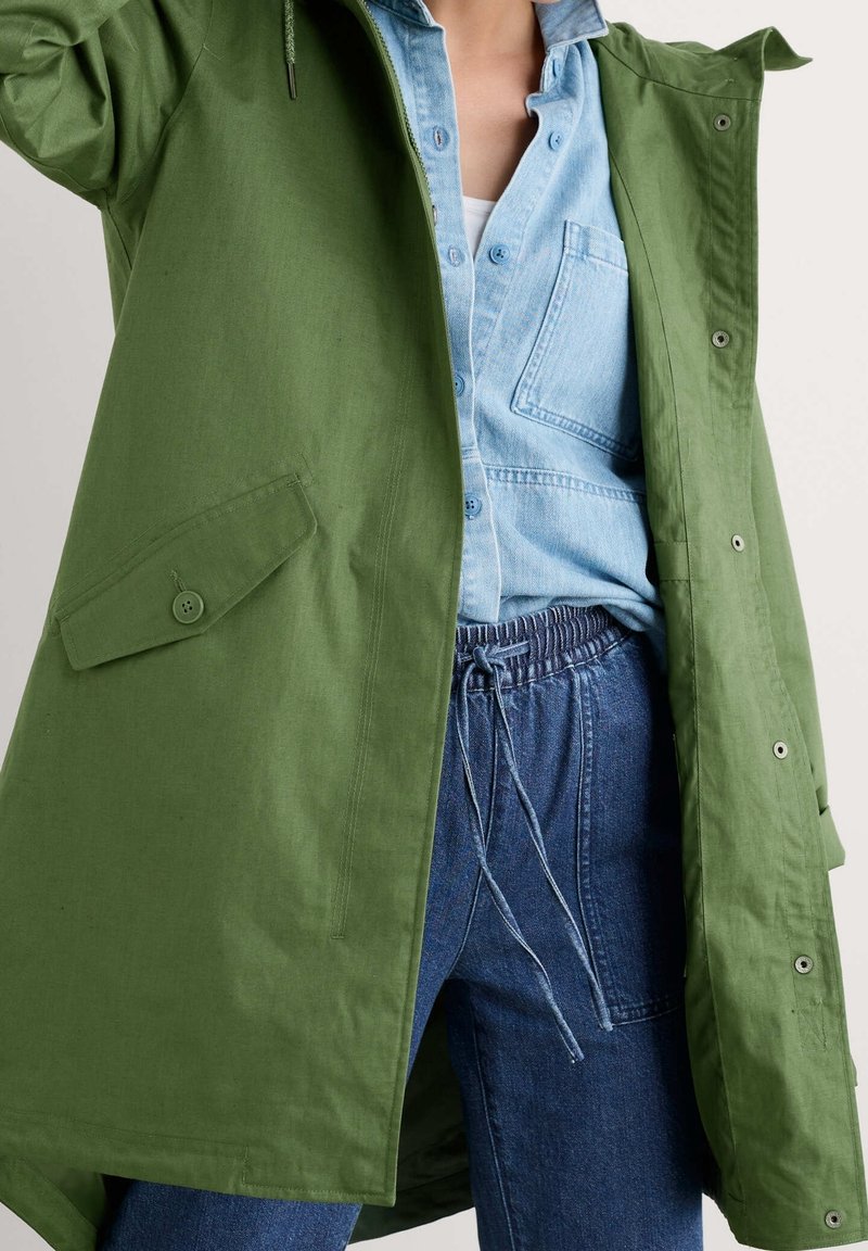 Grön parkas med huva och knappstängning framtill, med sidofickor, över en ljusblå denimskjorta och blå byxor med elastiskt midjeband.