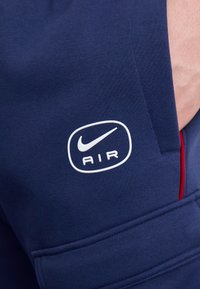 Granatowy materiał bluzy z gładką fakturą. Posiada białe logo Nike AIR w owalnym kształcie. Zawiera kontrastującą czerwoną kieszeń.