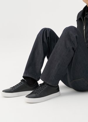 Personne assise avec les jambes pliées, portant un jean bleu foncé et des baskets basses noires avec des semelles blanches sur fond blanc.