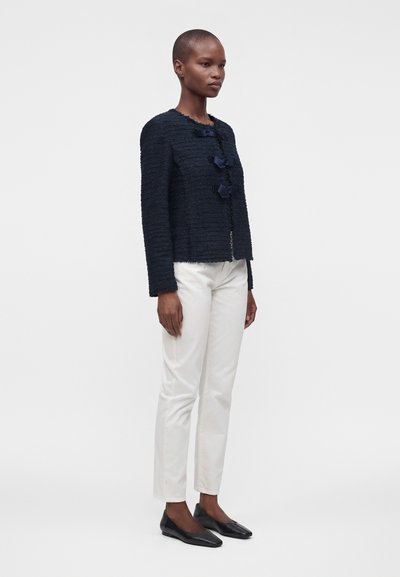 Femme debout de profil portant une veste texturée bleu marine avec des détails nœud, un pantalon blanc et des chaussures plates noires sur un fond blanc uni.