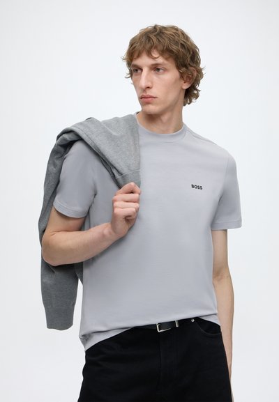 Jeune homme aux cheveux bouclés portant un t-shirt BOSS gris clair, tenant un pull gris sur l'épaule, debout devant un fond uni.