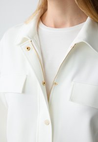 Wit gekraagde blazer met een gouden rits en knoopdetails, met een soepele textuur en twee kleine borstzakken.