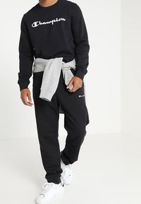 Svart Champion-hoodie med vitt logotyp, grå sweatshirt knuten i midjan, svarta joggers och vita sneakers. Avslappnad atletisk outfit.