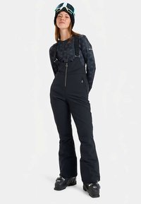 Kvinna i svarta skidbyxor, mönstrad långärmad tröja, svarta skidstövlar och skidglasögon på huvudet, stående mot en vit bakgrund.