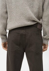 Jeans de mezclilla marrón con una textura suave, que cuentan con dos bolsillos traseros y una cinturilla con un detalle de cuero marrón. Suéter gris acanalado visible en la parte superior.