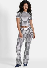 Mujer de pie con una camiseta de rayas horizontales en azul marino y blanco de manga corta y pantalones acampanados a juego, luciendo zapatillas azul marino sobre fondo blanco.
