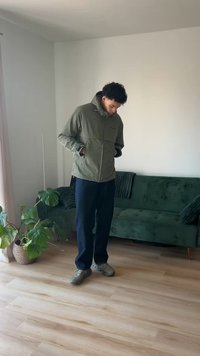 Oljčno zelena jakna s kapuco, z zadrgo spredaj in žepi. Kombinirana s temnimi hlačami in sivimi čevlji. Vidna lesena tla in zelena kavč.