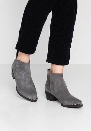 Nilkkapituiset harmaat mokkanahkasaappaat, joissa on paksu korko ja metallinhohtoiset varvasosat, yhdistetty mustiin polvipitkiin corduroshousuihin.