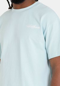 Camiseta de algodón de color azul claro con mangas cortas, que presenta un pequeño logotipo en cursiva blanca en la parte izquierda del pecho. Ajuste suave y relajado.
