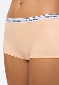 Κοντινό πλάνο ενός ατόμου που φοράει αντρικό εσώρουχο boyshort σε ροδακινί χρώμα της Calvin Klein με λευκή μέση που φέρει το όνομα της μάρκας σε μαύρο κείμενο.