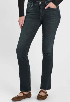 Vrouw draagt donkere slim-fit jeans, zwarte top met lange mouwen en bruine Mary Jane schoenen, staat met één hand in de zak.