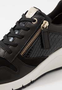 Černá sportovní obuv s hladkou kůží, texturovanými lakovanými akcenty, zlatým zipem a bílou podrážkou se vrstveným designem.