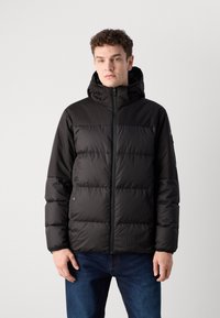 Zwarte pufferjack met een ritssluiting aan de voorkant en een capuchon. Heeft een gestikte design, zijzakken en geribbelde manchetten. Draagt over een blauwe spijkerbroek.