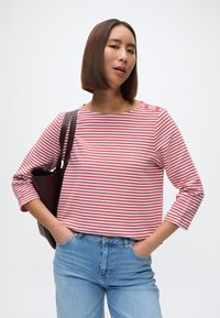 Λωρίδες κόκκινου και λευκού, μπλούζα τριών τετάρτων με στρογγυλή λαιμόκοψη και λεπτομέρεια κουμπιού. Συνδυασμένη με ανοιχτόχρωμο μπλε τζιν και σκούρη τσάντα.