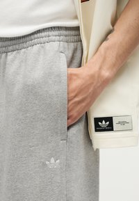 Χέρι που ακουμπά στην τσέπη γκρι φόρμας Adidas, φορώντας λευκό ribbed μπλουζάκι και κρατώντας μπεζ μπουφάν μπάσκετ Adidas με λογότυπο.