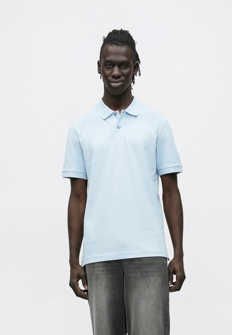 Mladý muž stojaci, oblečený v svetlomodrej polo košeli a vybledlých sivých džínsoch, na bielom neutrálnom pozadí.