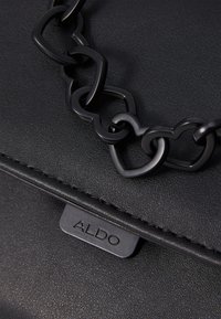 Schwarze Kunstleder-Handtasche mit einem herzförmigen Kettenriemen und geprägtem Logo am Verschluss. Strukturierte Oberfläche mit sauberen Nähten.