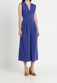 Jumpsuit sans manches bleu royal avec des jambes larges et un décolleté en V, porté avec des chaussures à talons hauts marron sur un fond neutre.