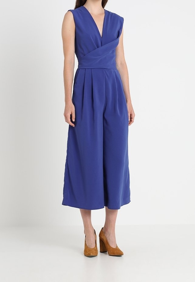Jumpsuit sans manches bleu royal avec des jambes larges et un décolleté en V, porté avec des chaussures à talons hauts marron sur un fond neutre.