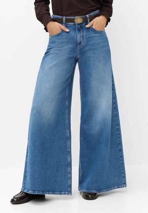 Person iført vidbenede blå jeans med et brunt bælte, mørk langærmet top og sorte sko, hænder i lommerne, stående på en hvid baggrund.