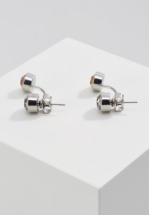Boucles d'oreilles clous en argent avec double sertissage rond, une pierre précieuse claire et une ambre, exposées sur un piédestal blanc.