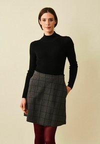 Kvinna som bär svart långärmad polotröja, grå kjol med svart rutmönster och burgunderröda strumpbyxor, stående mot en enfärgad beige bakgrund.