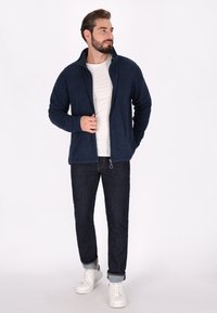 Giacca in pile blu navy con zip e colletto alto, indossata sopra una maglietta bianca, abbinata a jeans scuri e sneakers bianche. Vestibilità e tessuto casual.