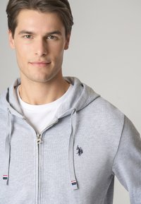 Felpa grigia con zip, realizzata in tessuto testurizzato. Presenta uno strato interno bianco e un logo ricamato sul petto, insieme a dettagli a righe sul cappuccio.