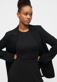 Μαύρο σακάκι με καθαρό σχέδιο, συνδυασμένο με μαύρο εφαρμοστό τοπ και φούστα. Λεία υφή, δομημένη εφαρμογή και ελάχιστες λεπτομέρειες.