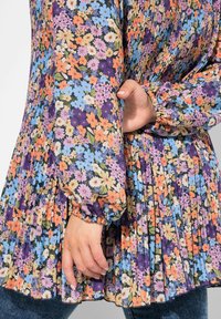 Květinové šaty s plisovaným designem, barevnými květinovými vzory v odstínech fialové, modré a oranžové, s nabíranými rukávy a měkkou tkaninou.